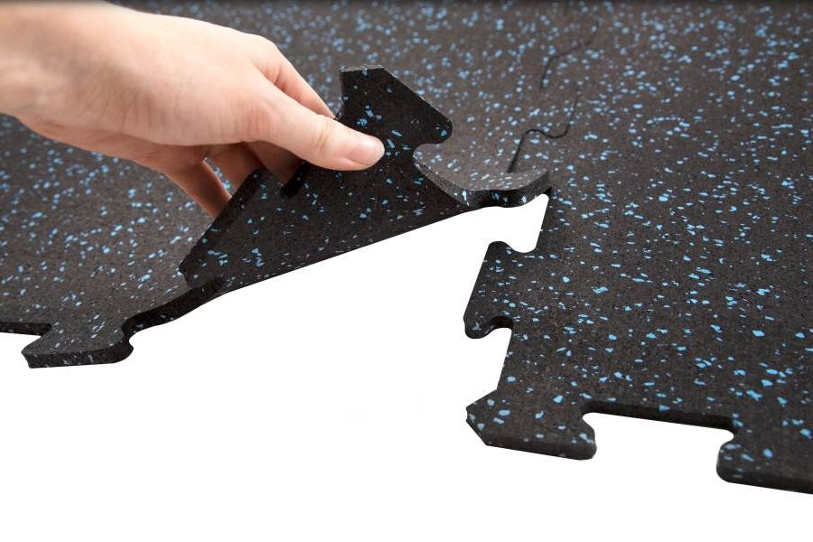 Hand holding black interlocking rubber gym floor tile with blue speckles, shown over white background