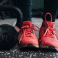 Black cast iron kettlebell and coral lace-up running shoes on speckled gray rubber gym flooring with person wearing dark legg
