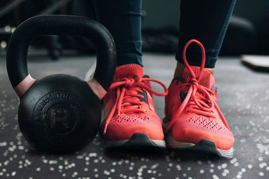 Black cast iron kettlebell and coral lace-up running shoes on speckled gray rubber gym flooring with person wearing dark legg