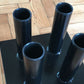 Black metal vertical barbell holder with five cylindrical tubes mounted on square base, displayed on wooden floor