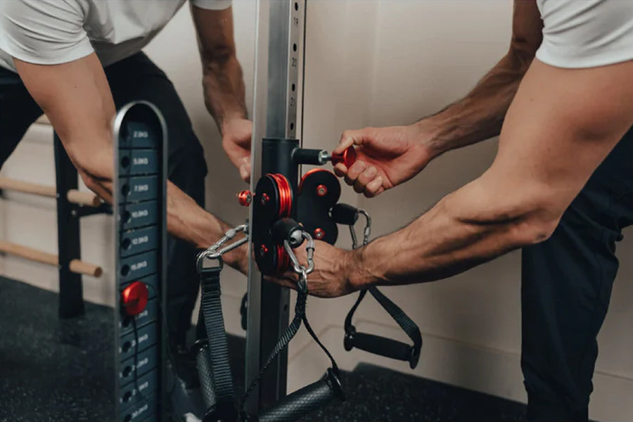 Two people assembling red and black cable pulley system on grey wall-mounted gym equipment with digital display panel