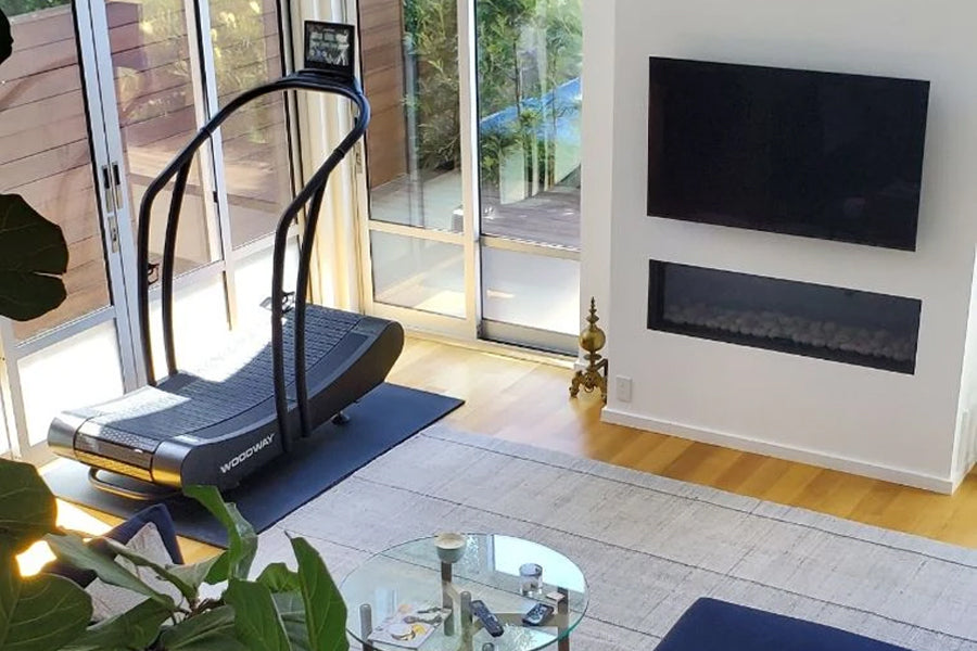 Woodway Curve Treadmill with black frame and blue mat in modern home gym setting with glass doors and wall-mounted TV