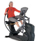 Woman in red jacket and gray capris using black and gray commercial recumbent elliptical machine on white background
