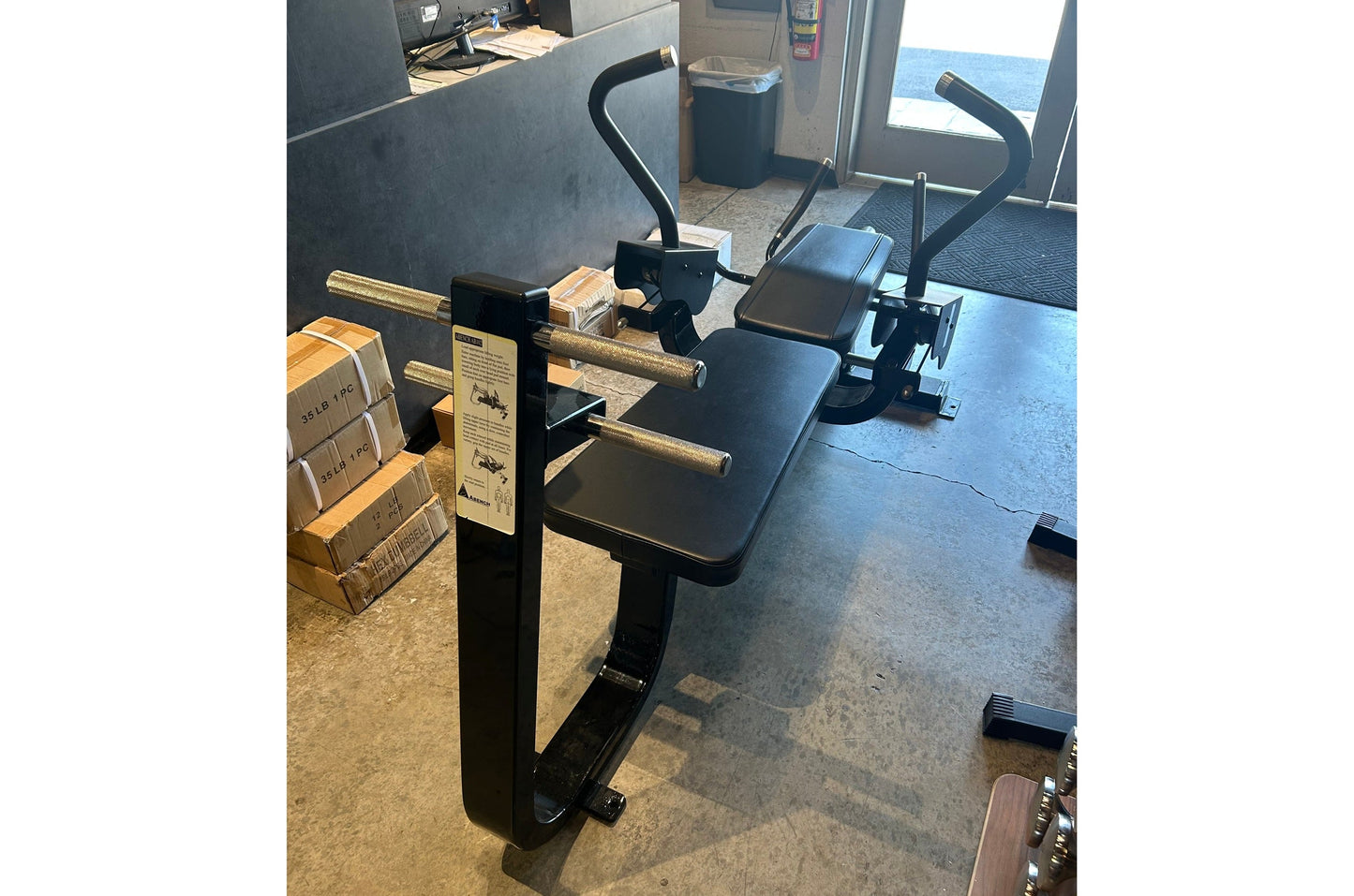 Black commercial abdominal bench with padded seat and backrest, gold metal frame, black handles, in gym setting with cardboar