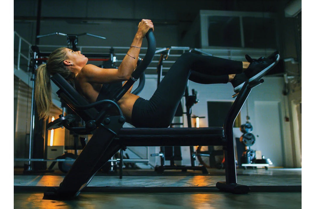 Woman using black commercial abdominal bench in gym setting, performing core exercise with legs extended