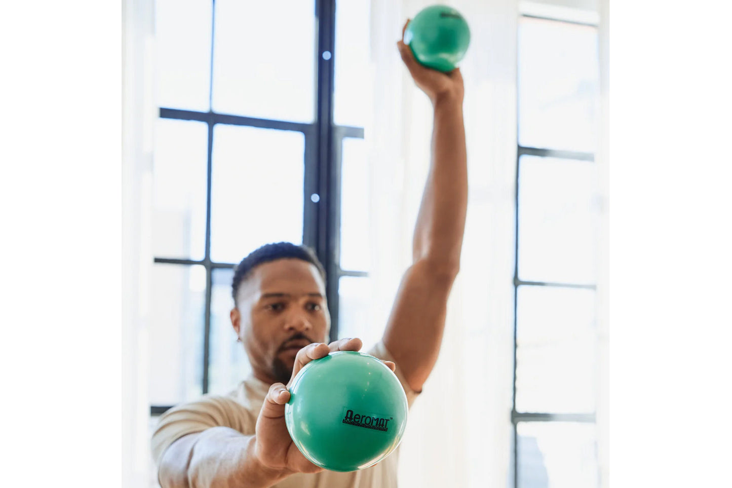 Man holding green Aeromat mini weight balls, one in each hand, demonstrating exercise use by bright window