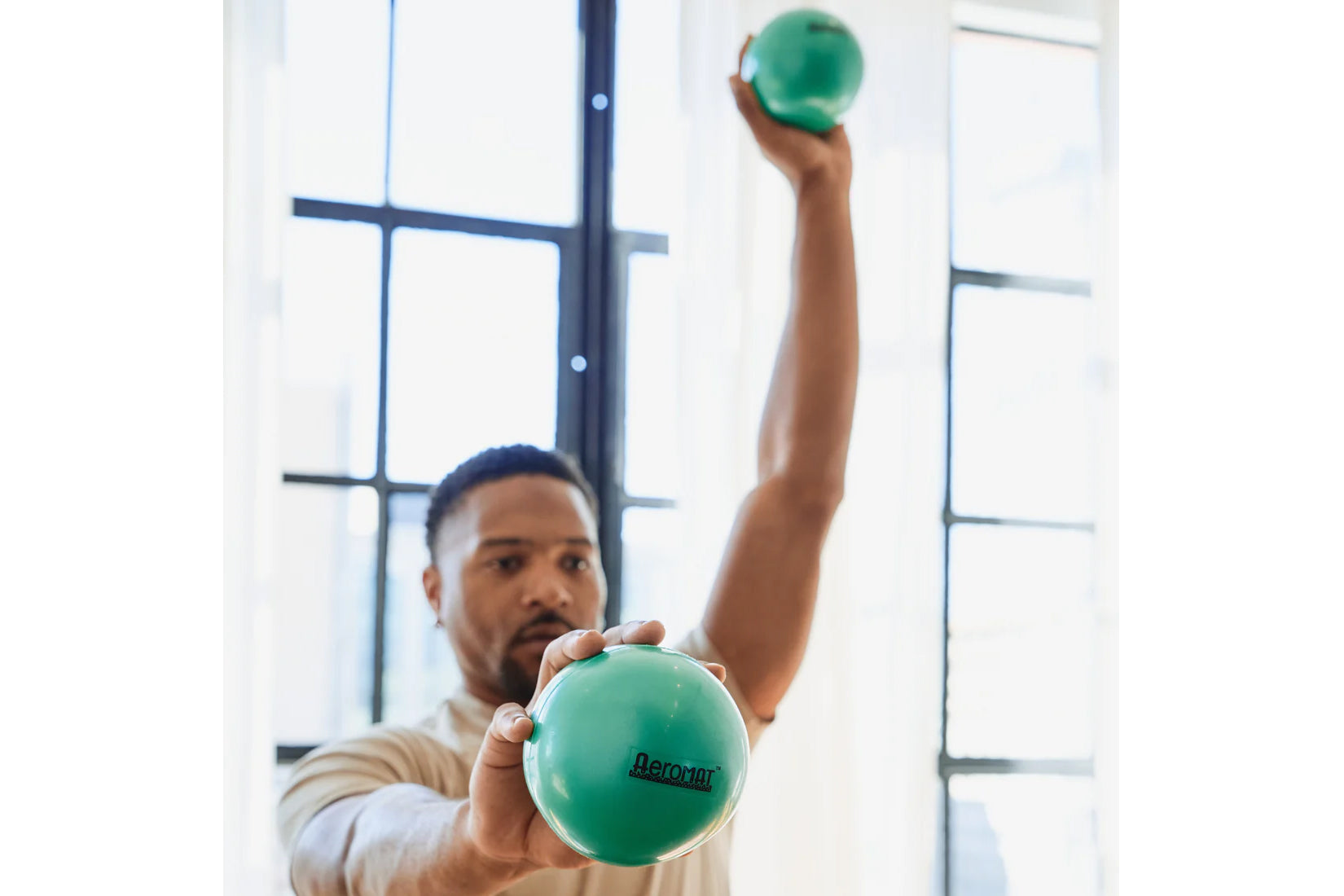 Man holding green Aeromat mini weight balls, one in each hand, demonstrating exercise use by bright window