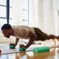 Man in beige shirt and olive shorts doing a plank exercise with green Aeromat Mini Weight Balls on yoga mat in bright studio