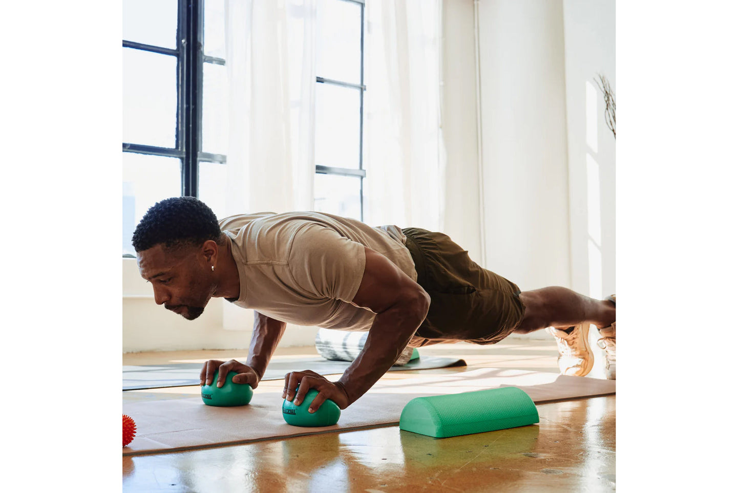 Man in beige shirt and olive shorts doing a plank exercise with green Aeromat Mini Weight Balls on yoga mat in bright studio