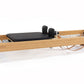 Natural wood pilates reformer with black padded carriage, footbar, and black foot blocks on white background