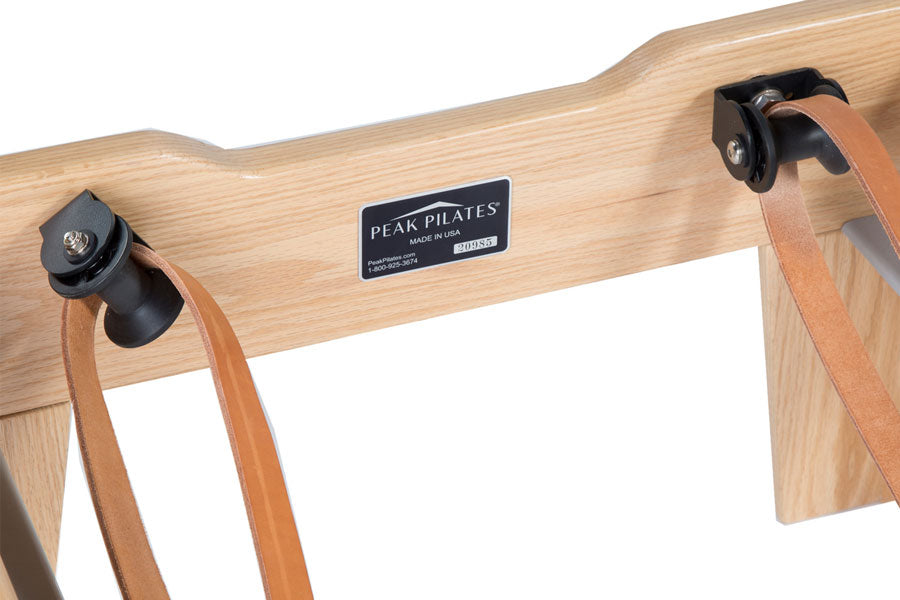 Natural wood Pilates reformer with Peak Pilates nameplate, tan leather straps, and black adjustment hardware on white backgro