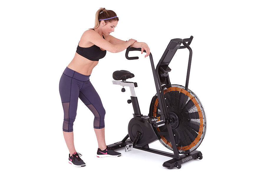 Woman in black sports bra and purple capri leggings using black and orange Octane Fitness AirdyneX exercise bike on white bac