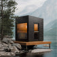Modern black wooden sauna with orange interior, glass door and window, mounted on wooden deck over mountain lake