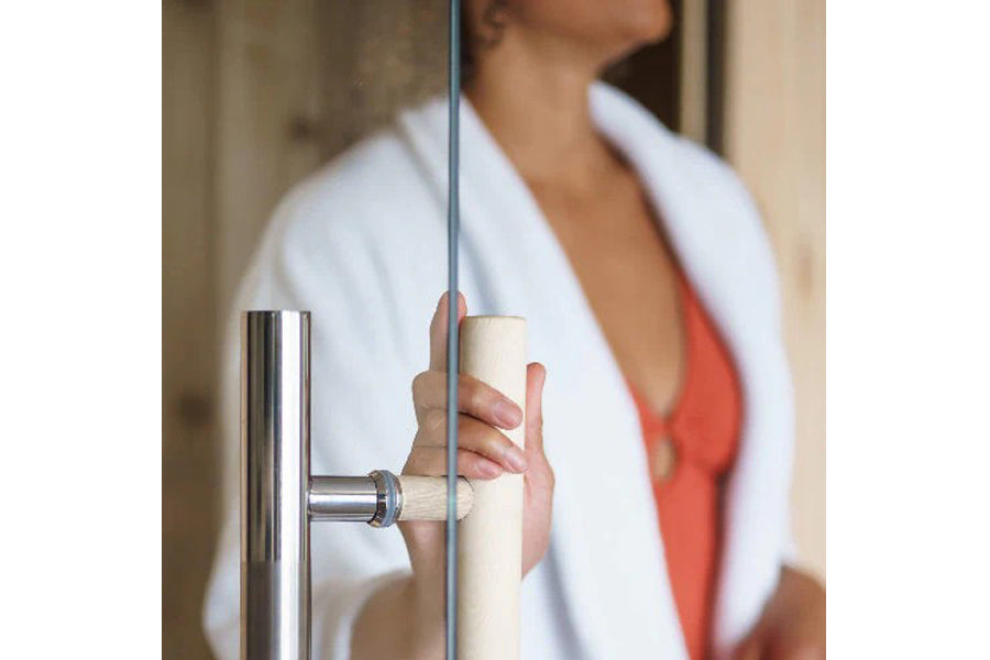 Model opening glass door of indoor sauna with white towel wrapped around shoulders and red swimsuit visible