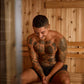 Man with tattooed torso sitting inside wooden sauna with light wood paneling
