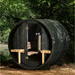 Black horizontal barrel sauna with wooden benches inside, open door showing interior, surrounded by green foliage