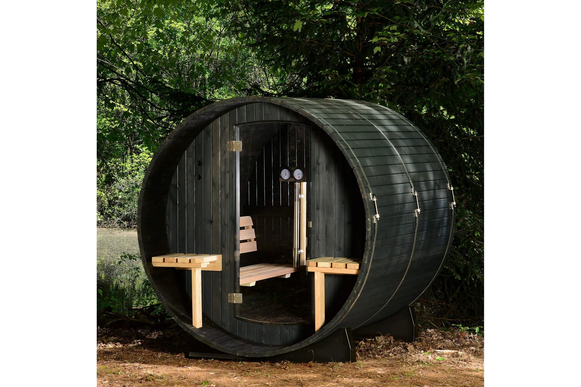 Black horizontal barrel sauna with wooden benches inside, open door showing interior, surrounded by green foliage