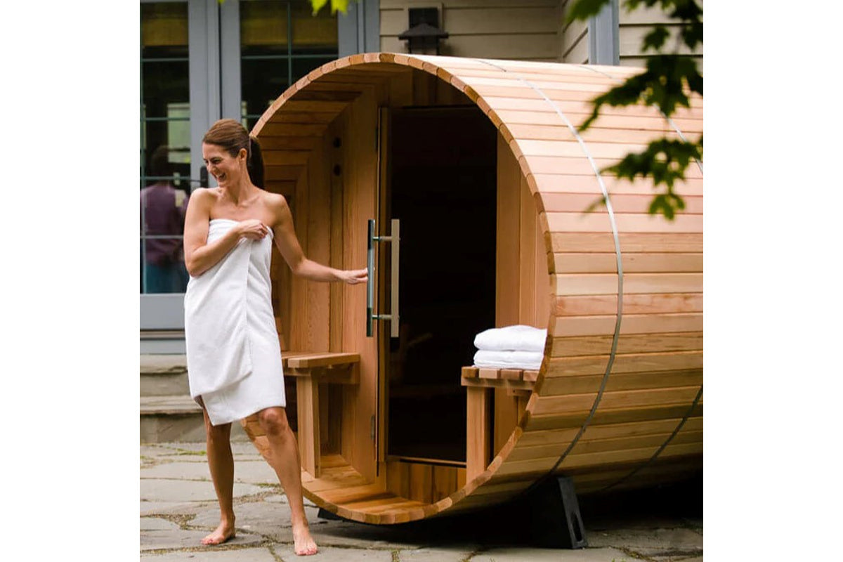 Woman in white towel standing at entrance of wooden barrel sauna with curved roof and metal bands