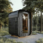Black wood-sided sauna with rounded corners and glass door, positioned on wooden deck in forest setting