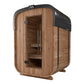 Compact wooden sauna with rounded top, cedar construction, dark metal roof, glass door, and bench seating visible inside, sho