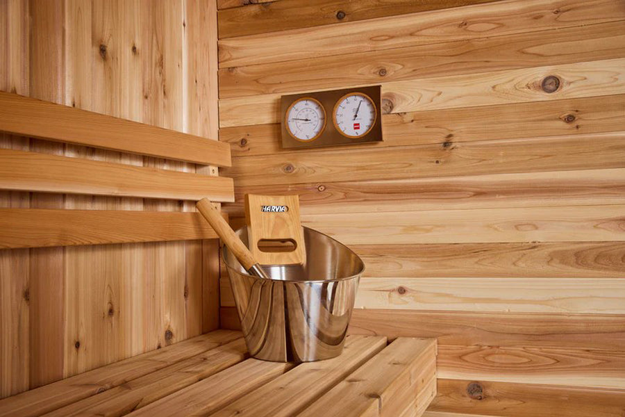 Cedar sauna interior with wooden bench, metal bucket with ladle, wooden control panel, and wall-mounted thermometer and hygro
