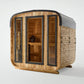 Cedar wood cube sauna with black metal frame, glass doors and side panels, featuring wooden interior bench, on white backgrou