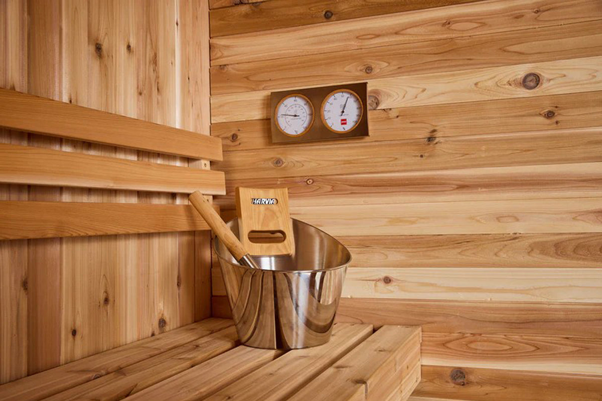Cedar sauna interior with wooden benches, metal water bucket with wooden ladle, and dual analog thermometer and hygrometer mo