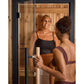 Woman in black ribbed crop top and tan bottoms smiling inside wooden sauna with glass door and cedar interior