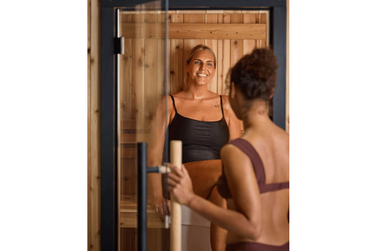 Woman in black ribbed crop top and tan bottoms smiling inside wooden sauna with glass door and cedar interior