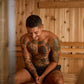 Shirtless man with full-body tattoos sitting inside wooden sauna with natural pine walls and benches