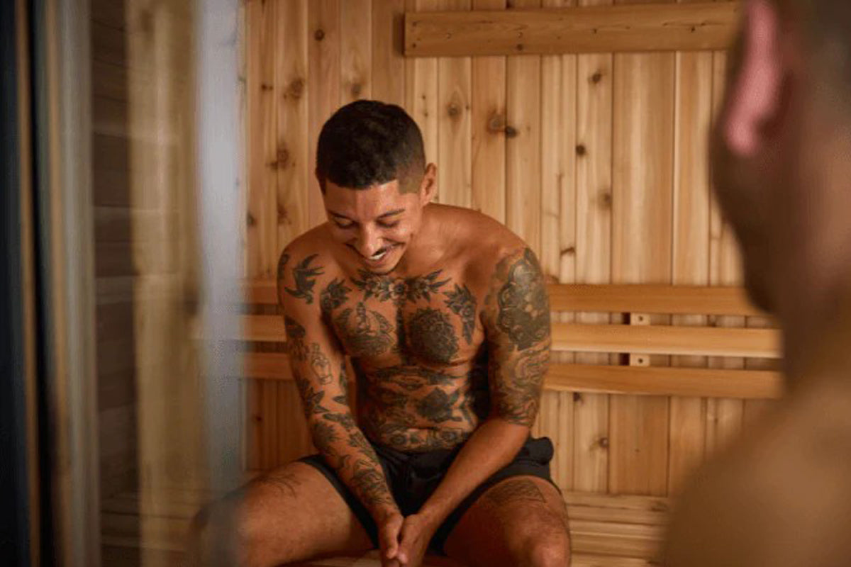 Shirtless man with full-body tattoos sitting inside wooden sauna with natural pine walls and benches