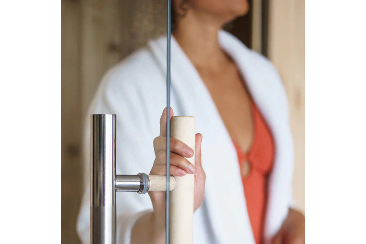 Woman opening glass sauna door with cream-colored handle and stainless steel frame, wearing white robe over coral top
