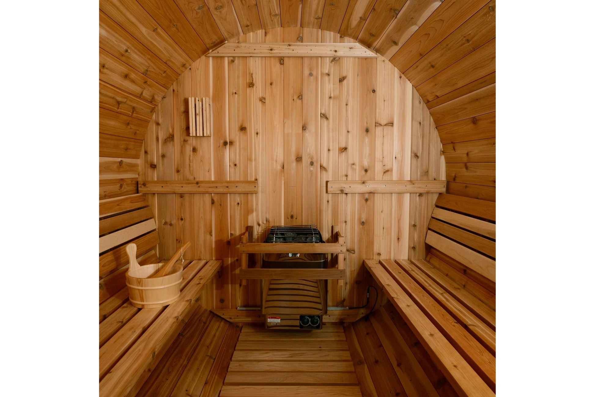 Cedar barrel sauna interior with curved wooden walls, built-in benches on both sides, heater unit at back, wooden bucket, and