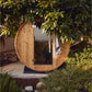 Round wooden barrel sauna with glass door and metal hardware, surrounded by green foliage in outdoor garden setting
