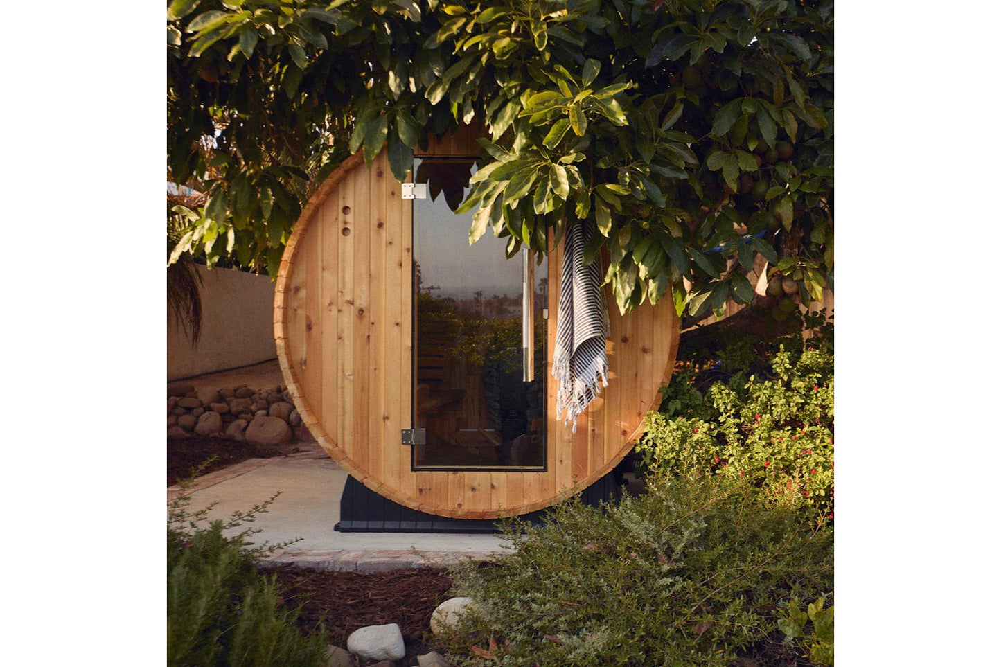 Round wooden barrel sauna with glass door and metal hardware, surrounded by green foliage in outdoor garden setting