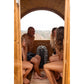 Family of four sitting inside wooden barrel sauna with half-moon canopy, viewed through open door with countryside landscape 