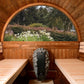Cedar barrel sauna interior with two wooden benches, black sauna stove with metal basket, arched window overlooking garden wi