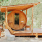 Cedar barrel sauna with round wooden door and window, mounted on wooden deck with pergola roof and slide entrance, surrounded