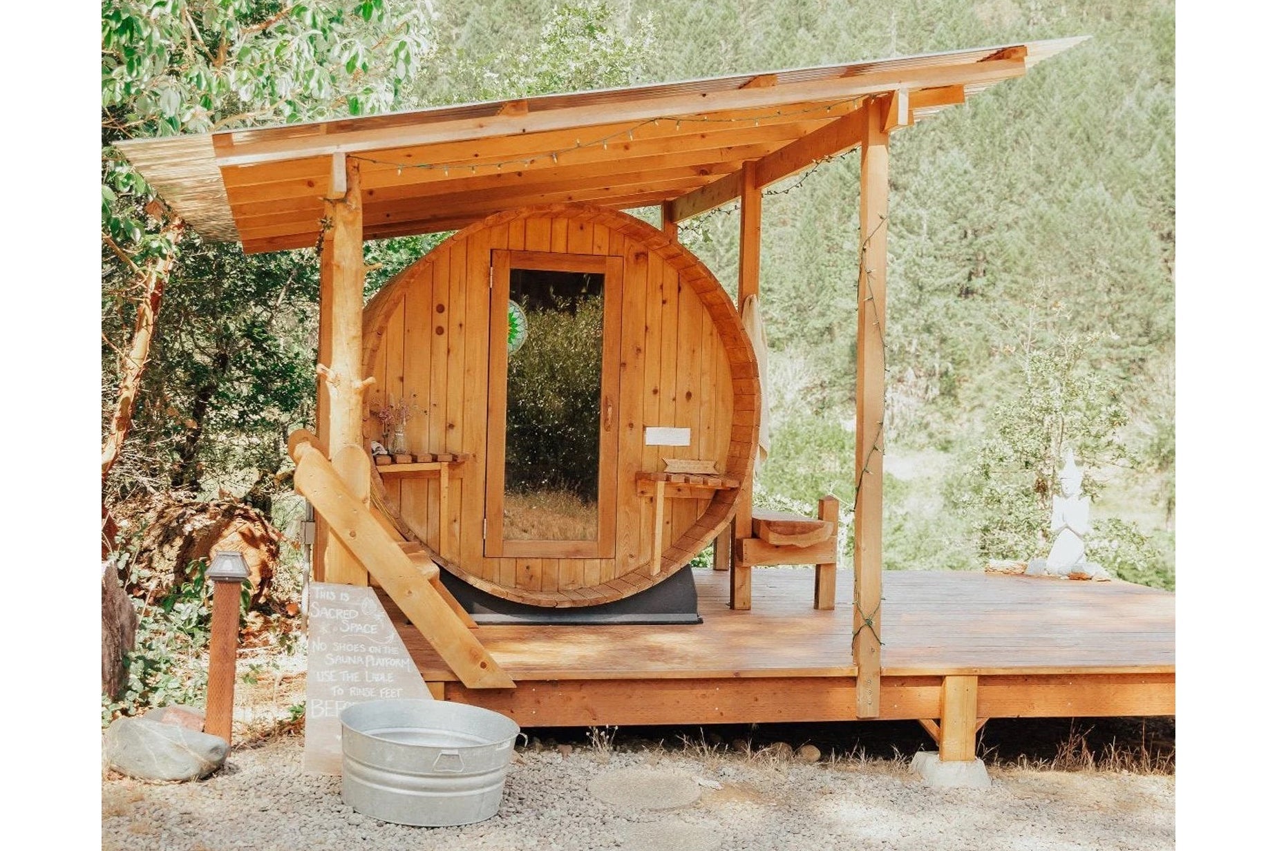Cedar barrel sauna with round wooden door and window, mounted on wooden deck with pergola roof and slide entrance, surrounded