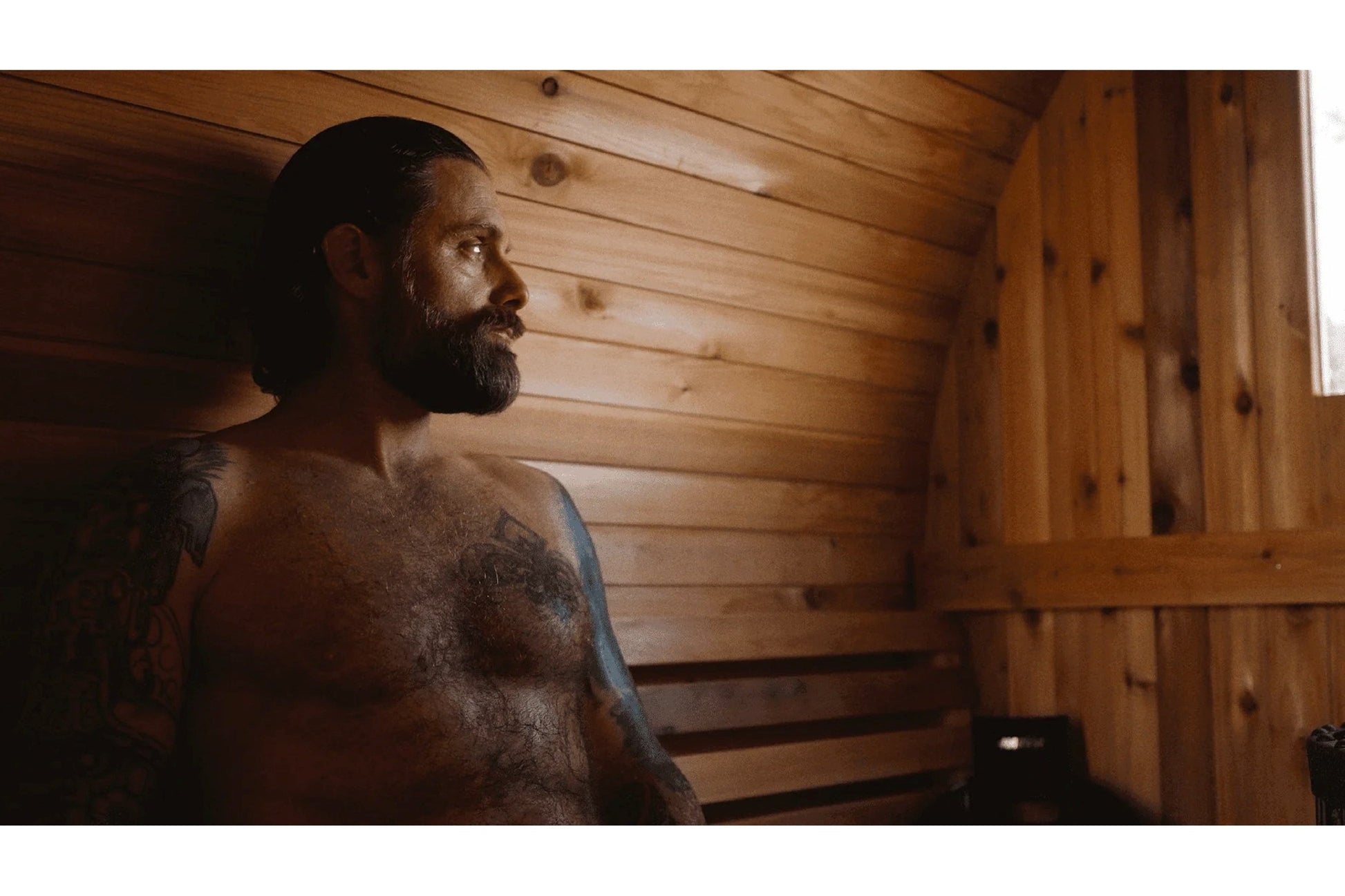 Man with beard and tattoos sitting shirtless inside wooden sauna with horizontal wooden benches