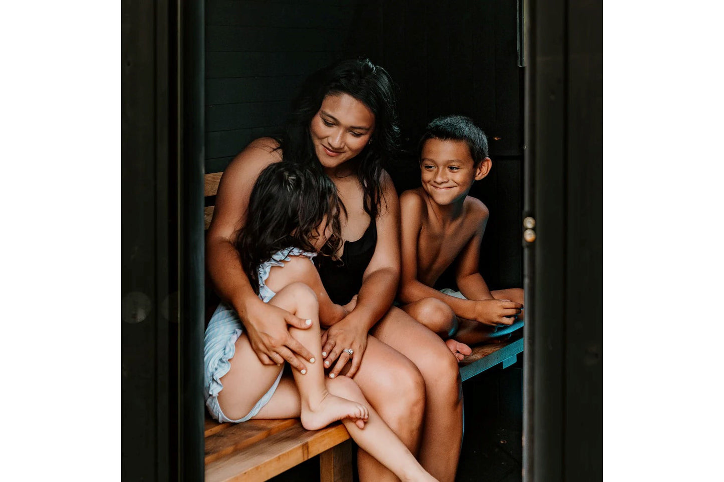 Woman and two children sitting together in wooden sauna barrel with black canopy, viewed through open door