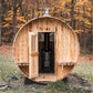Wooden barrel sauna with light natural wood exterior, open door revealing black interior stove, metal roof cap, set in autumn