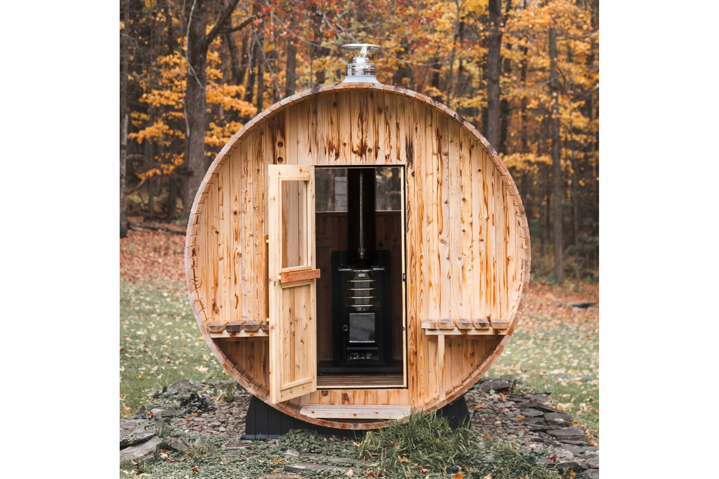 Wooden barrel sauna with light natural wood exterior, open door revealing black interior stove, metal roof cap, set in autumn
