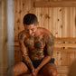 Man with full-sleeve tattoos sitting in wooden sauna interior with light wood paneling