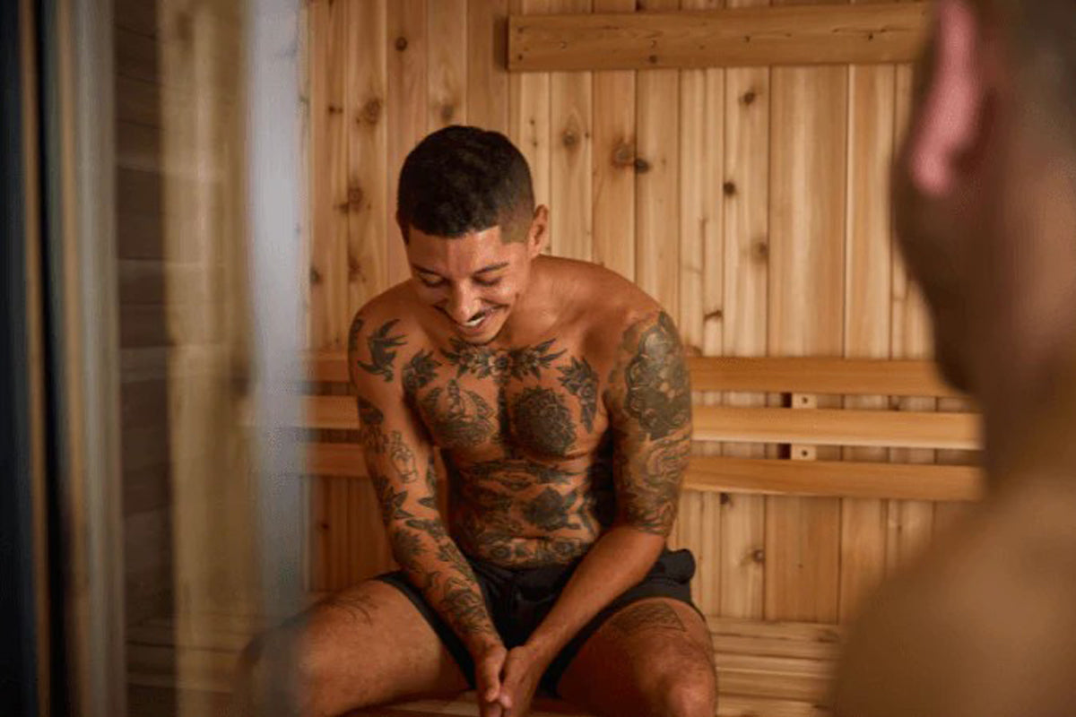 Man with full-sleeve tattoos sitting in wooden sauna interior with light wood paneling