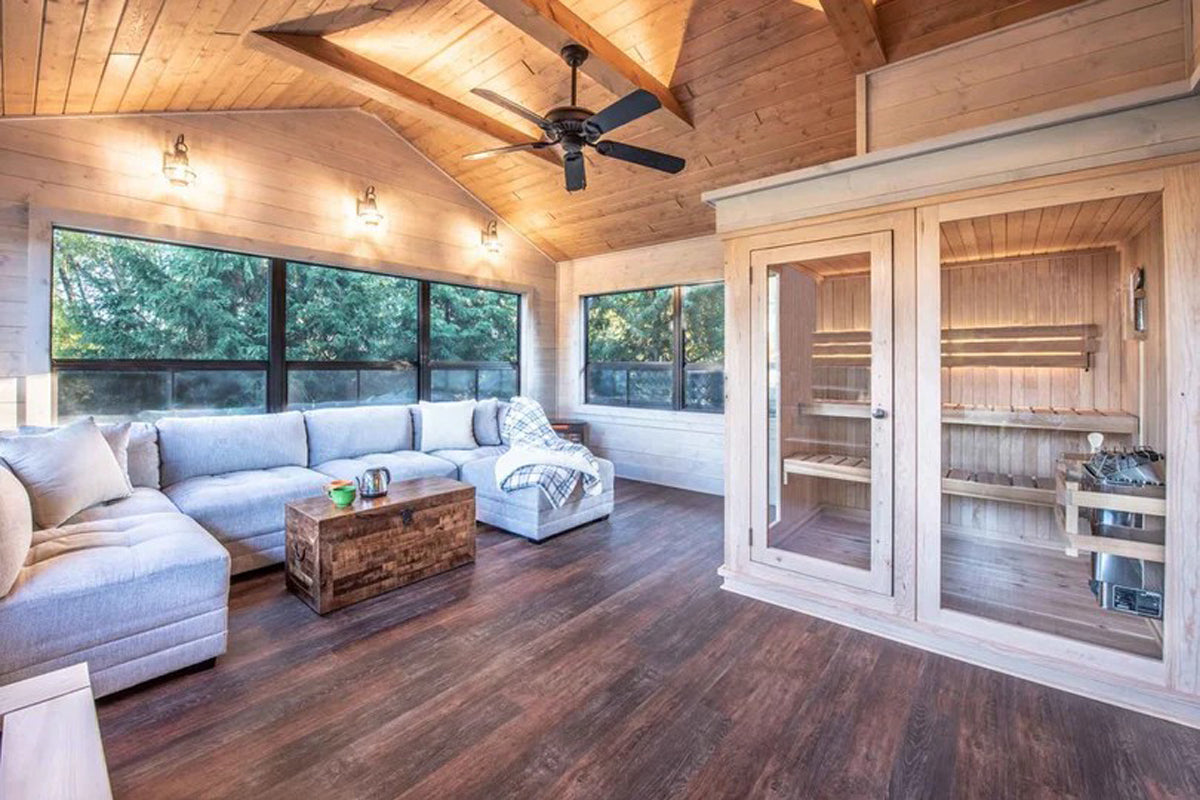 Modern sauna room with light wood interior, glass doors, and cedar benches visible inside adjacent to a living area with blue