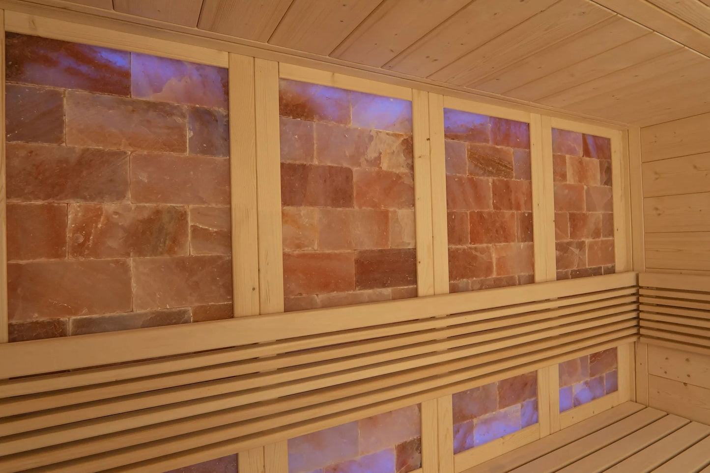 Interior of wooden sauna with light wood paneling, red salt brick walls backlit with blue and purple lighting, and wooden ben
