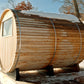 Cylindrical wooden barrel sauna with arched door and window, horizontal log construction, metal bands, and snow-covered roof 