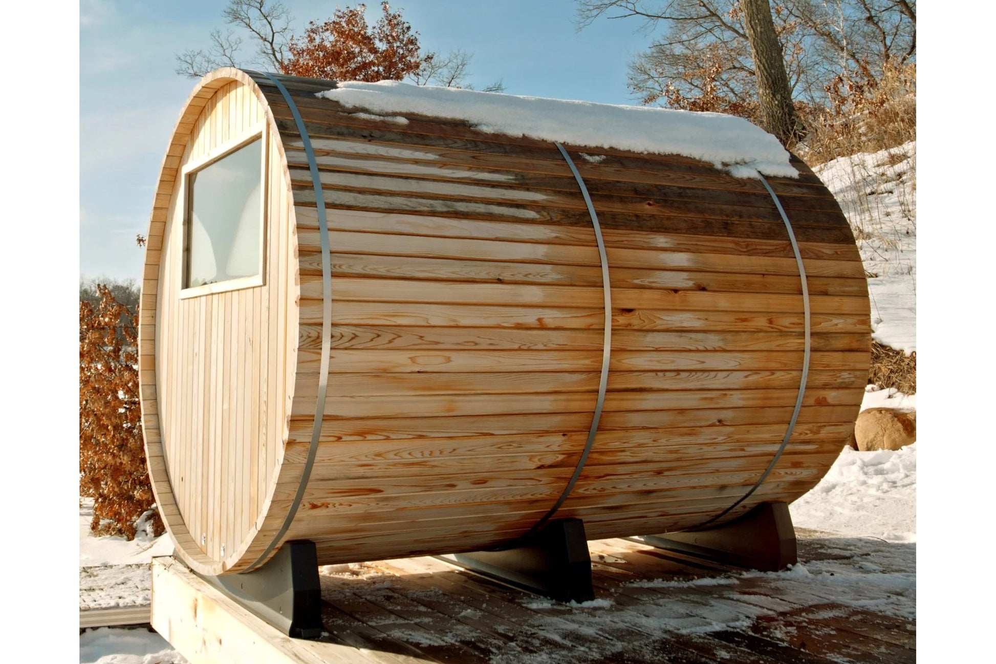 Cylindrical wooden barrel sauna with arched door and window, horizontal log construction, metal bands, and snow-covered roof 