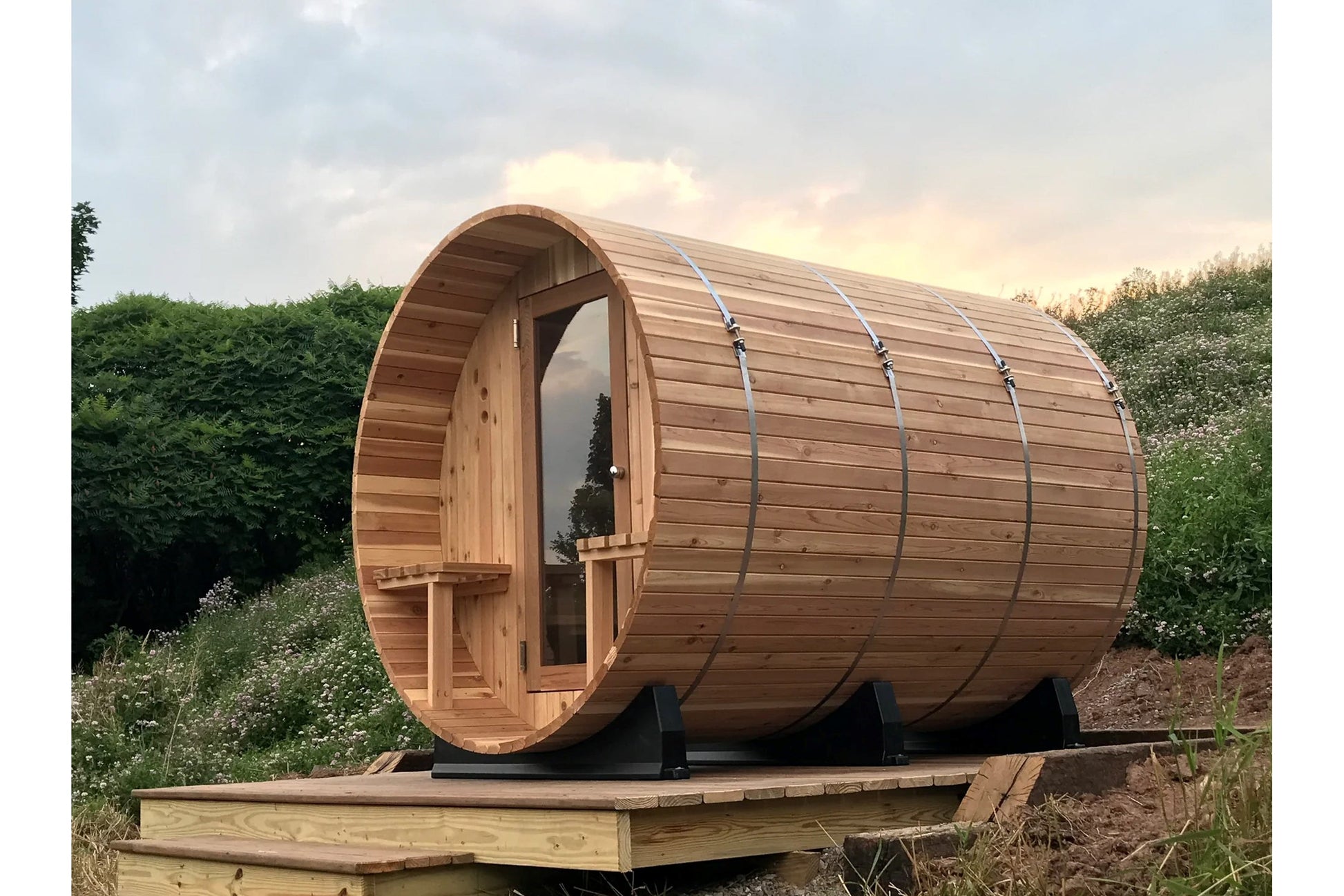 Barrel sauna with natural wood construction and curved cylindrical design, mounted on wooden platform in garden setting